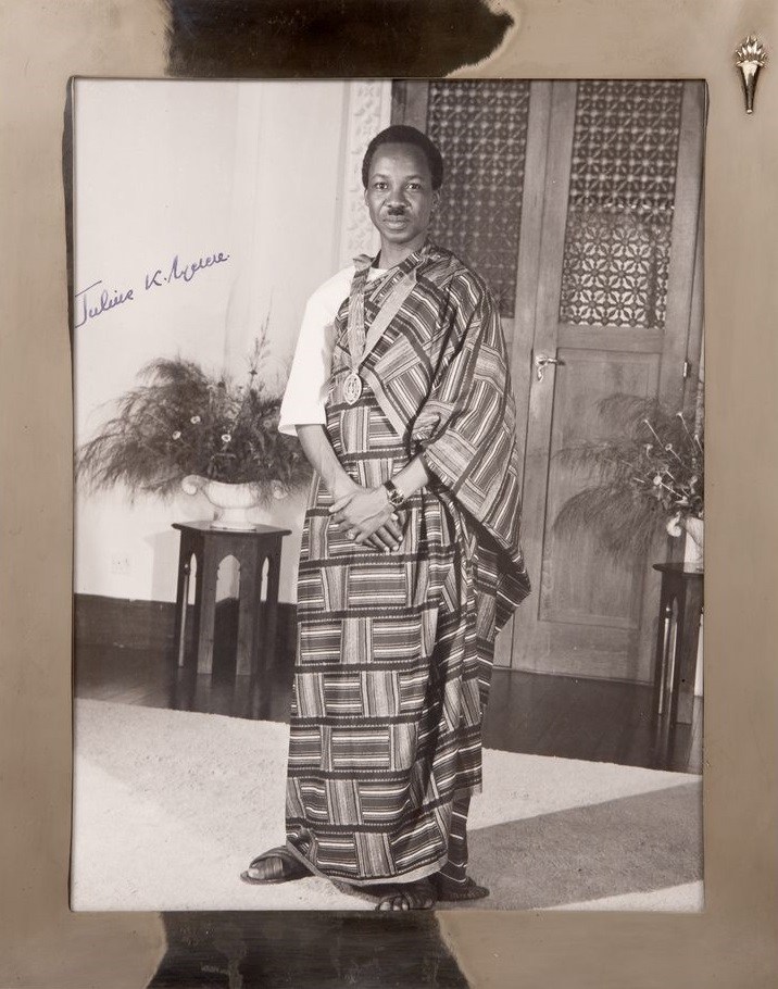 A rare portrait of   Julius K. Nyerere, President of Tanganyika (Tanzania). He is wearing traditional African dress with a medal around his neck. He stands in front of an ornately carved wood door flanked by two floral arrangements. The photograph is in a silver frame. Historical Note: This photograph was presented to President John F. Kennedy by Julius K. Nyerere, Prime Minister of Tanganyika (1961), President of Tanzania (1964-1985), during his state visit to the White House July 15, 1963. 